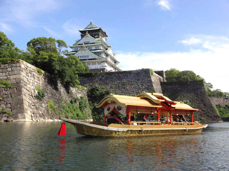 A traditional-style boat with a golden roof on a river near a castle with white walls and tiered roofs surrounded by trees.