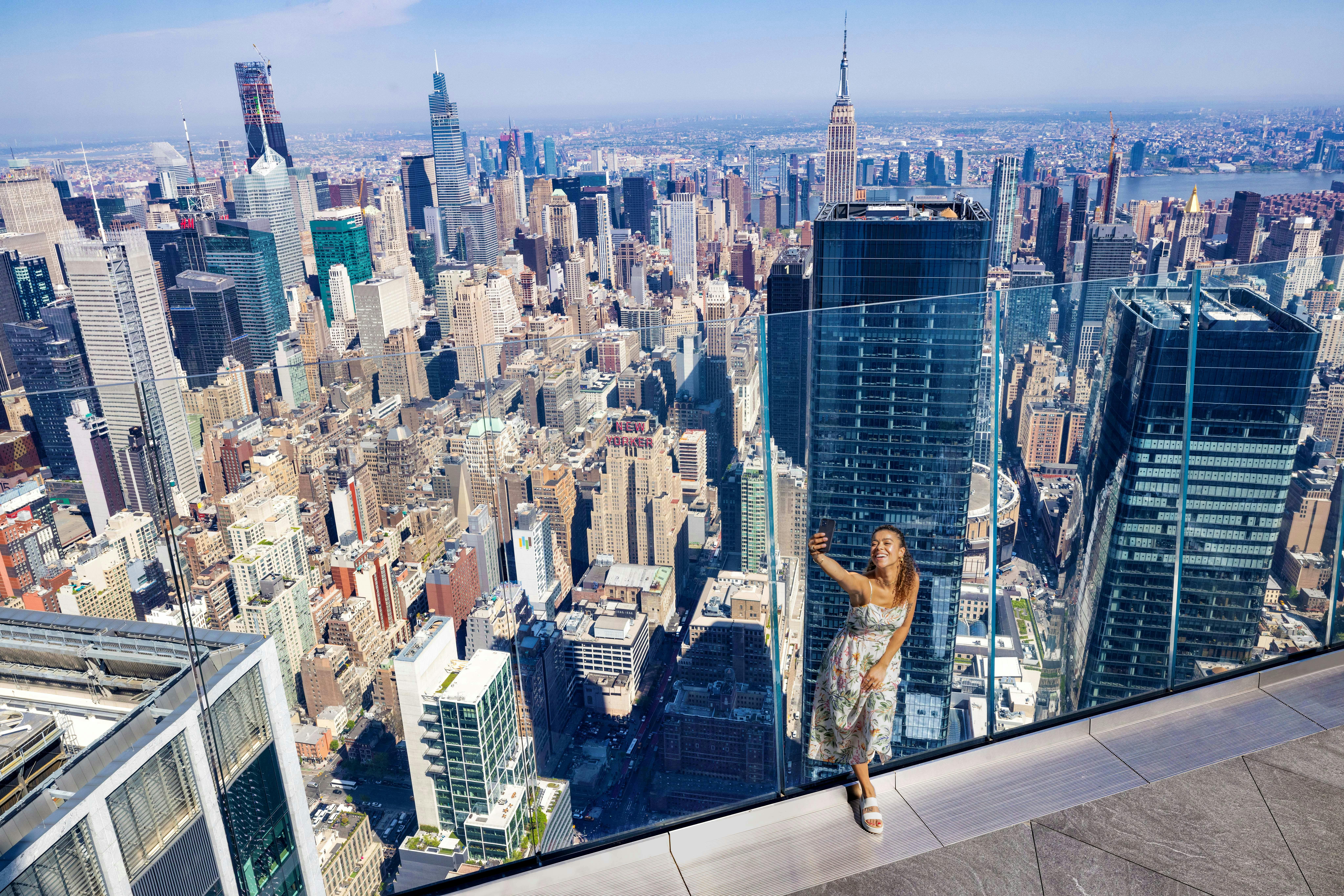 A woman takes a selfie on a rooftop observation deck with a view of a city skyline, including the Empire State Building.