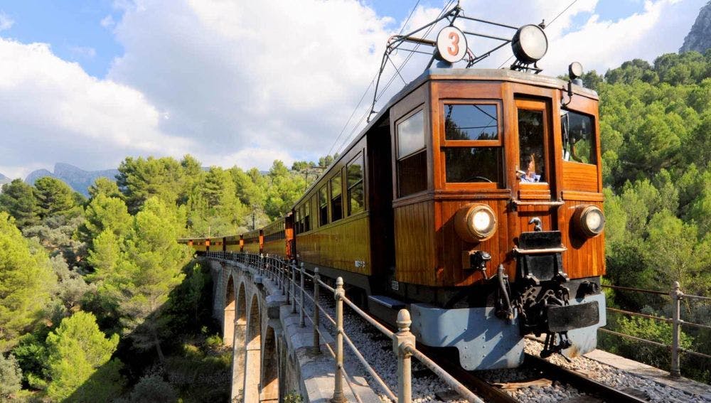 A vintage wooden train number 3 crosses an arched stone bridge, surrounded by lush green trees under a partly cloudy sky.