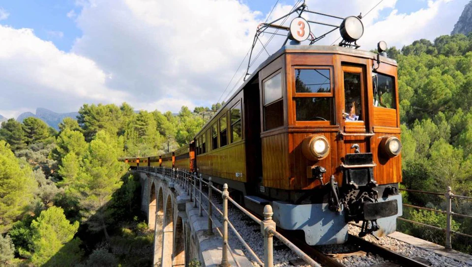 A vintage wooden train number 3 crosses an arched stone bridge, surrounded by lush green trees under a partly cloudy sky.
