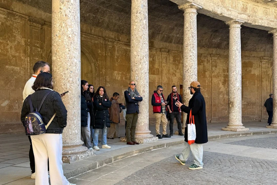 A group of people standing and listening to a guide in an outdoor colonnade with ornate stone pillars and an arched ceiling.