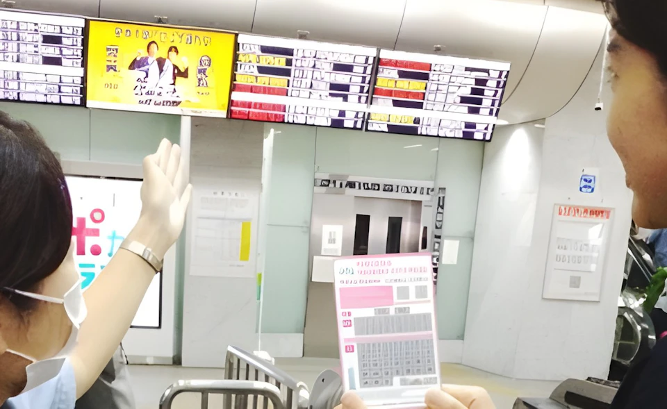 Two individuals at a ticket gate in a train station, one holding a ticket and the other pointing at an information board.