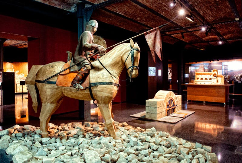 A museum exhibit featuring a knight in armor on a wooden horse, surrounded by rocks, with period artifacts displayed behind.