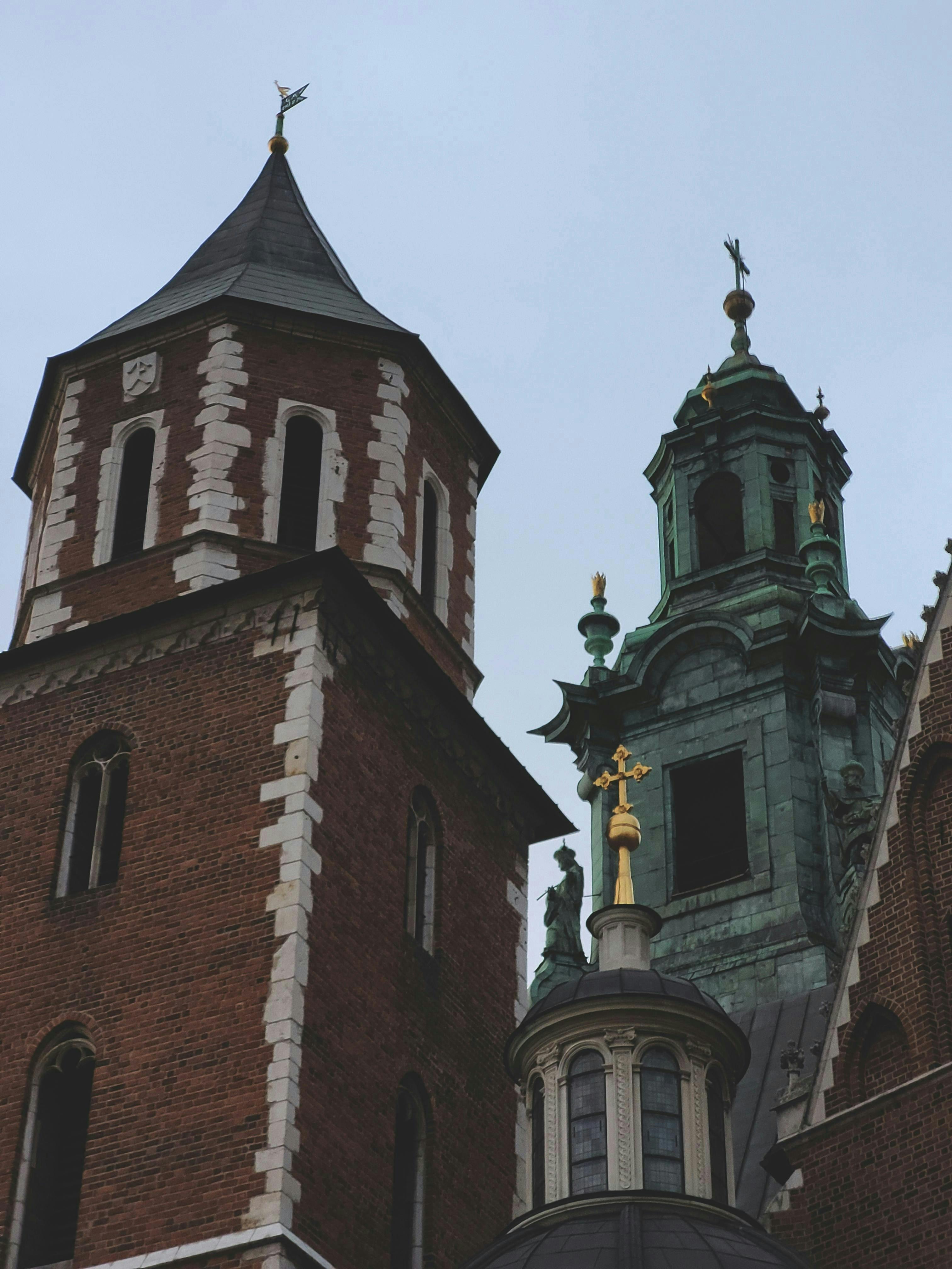 Two historic church towers with different architectural styles against a cloudy sky, featuring a cross on one spire.