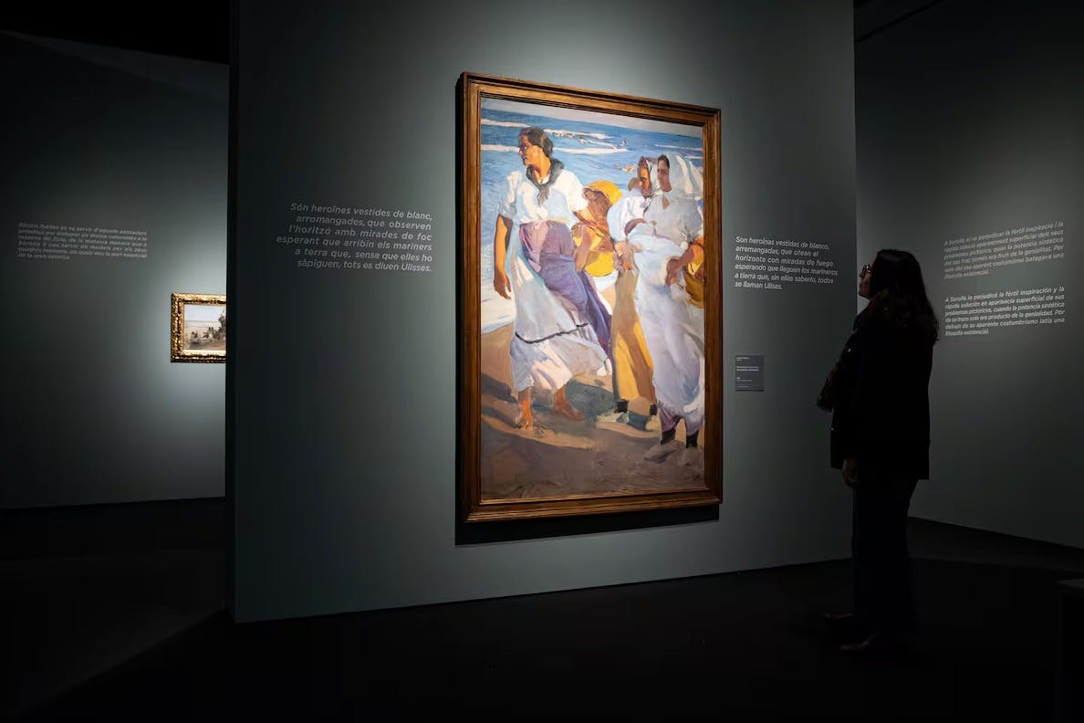 Картина, выставленная в музее, изображающая женщин на пляже, на которую смотрит человек, стоящий неподалеку в тускло освещенной комнате.