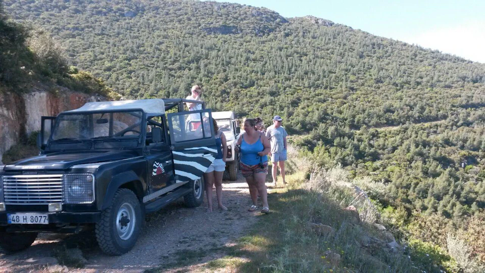 People standing near off-road vehicles on a dirt path surrounded by dense green forest and hills.