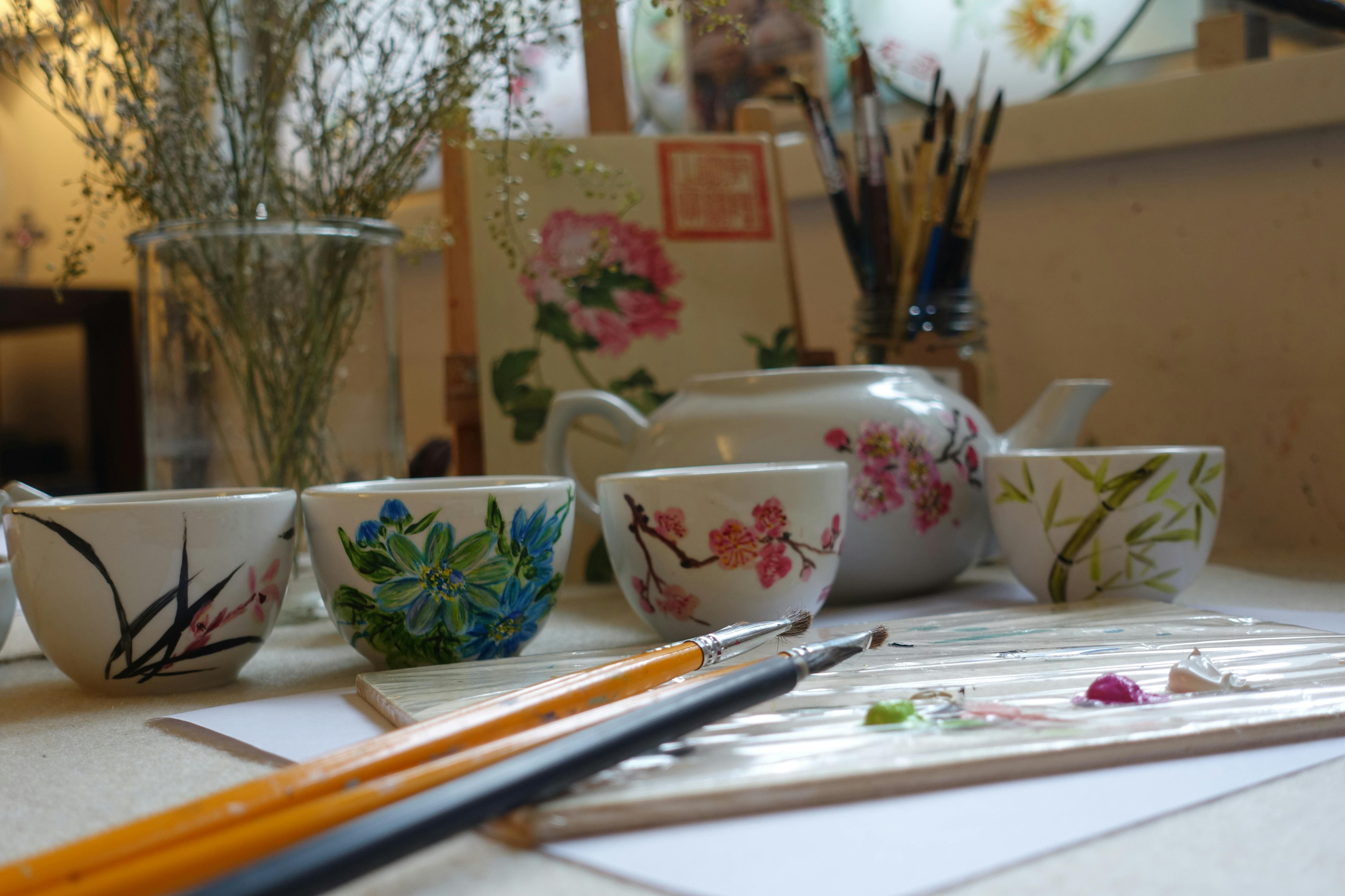 画笔、花卉绘制的茶杯、配套的茶壶和艺术工作室桌上的植物艺术品。
