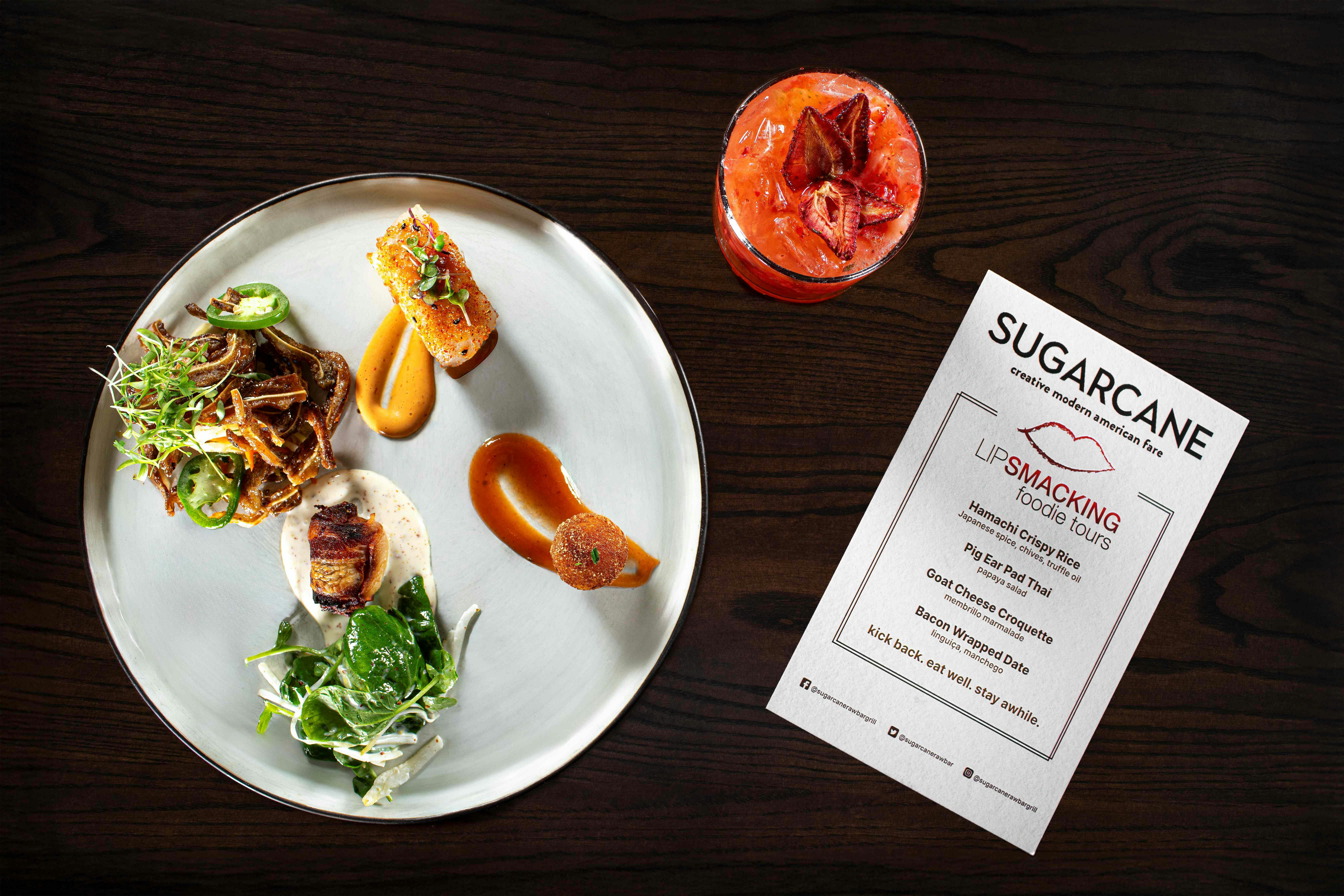 Ένα πιάτο γκουρμέ φαγητό με διάφορα είδη, ένα κόκκινο ρόφημα και ένα μενού σε ένα σκούρο ξύλινο τραπέζι.