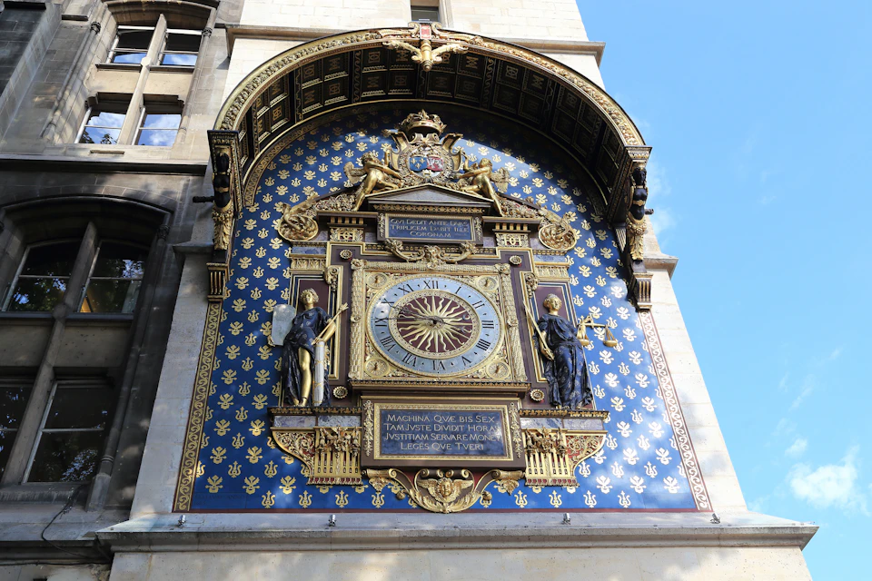 Ornate clock on a building facade with intricate details, two statues, a blue background with gold fleur-de-lis patterns.