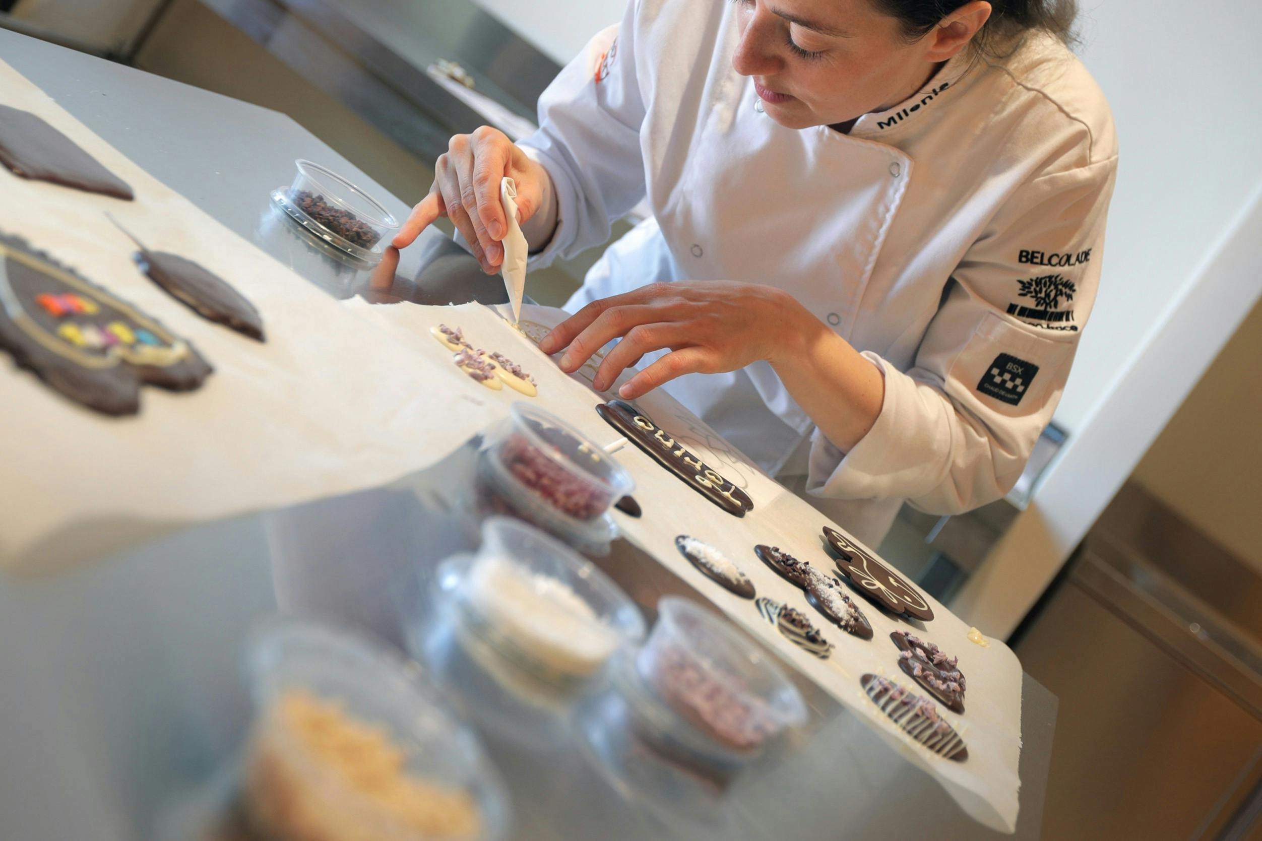 A pastry chef in a white uniform decorates chocolate pieces on parchment paper, with bowls of ingredients on the table.