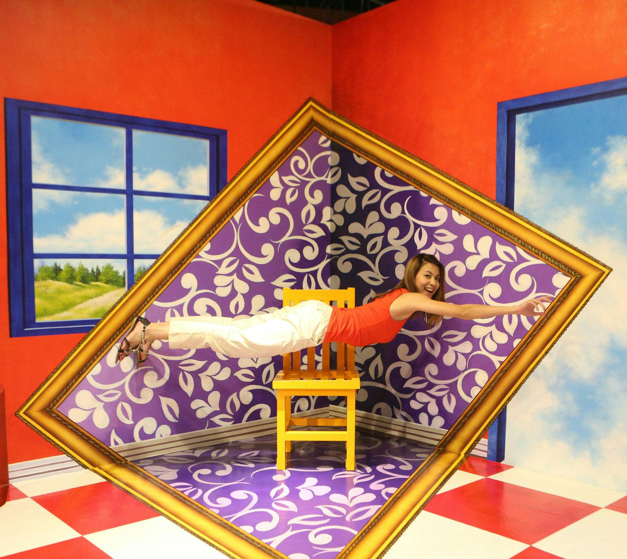 A woman is posing horizontally on a chair framed diagonally, with a vibrant, patterned background and checkered floor.