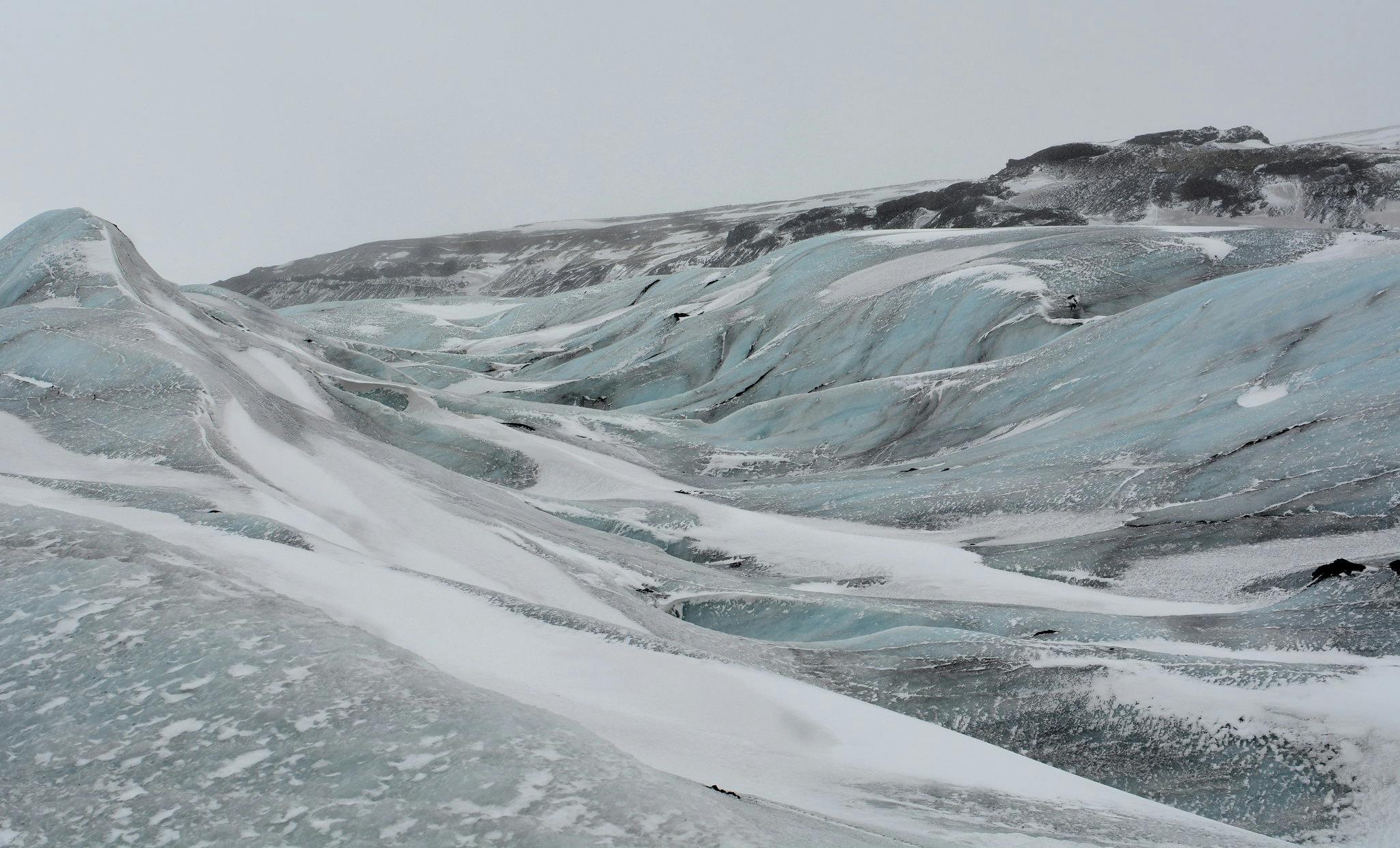 Besneeuwde gletsjer met blauwe ijsformaties onder een bewolkte hemel. Ruig terrein en vage bergtoppen in de verte.