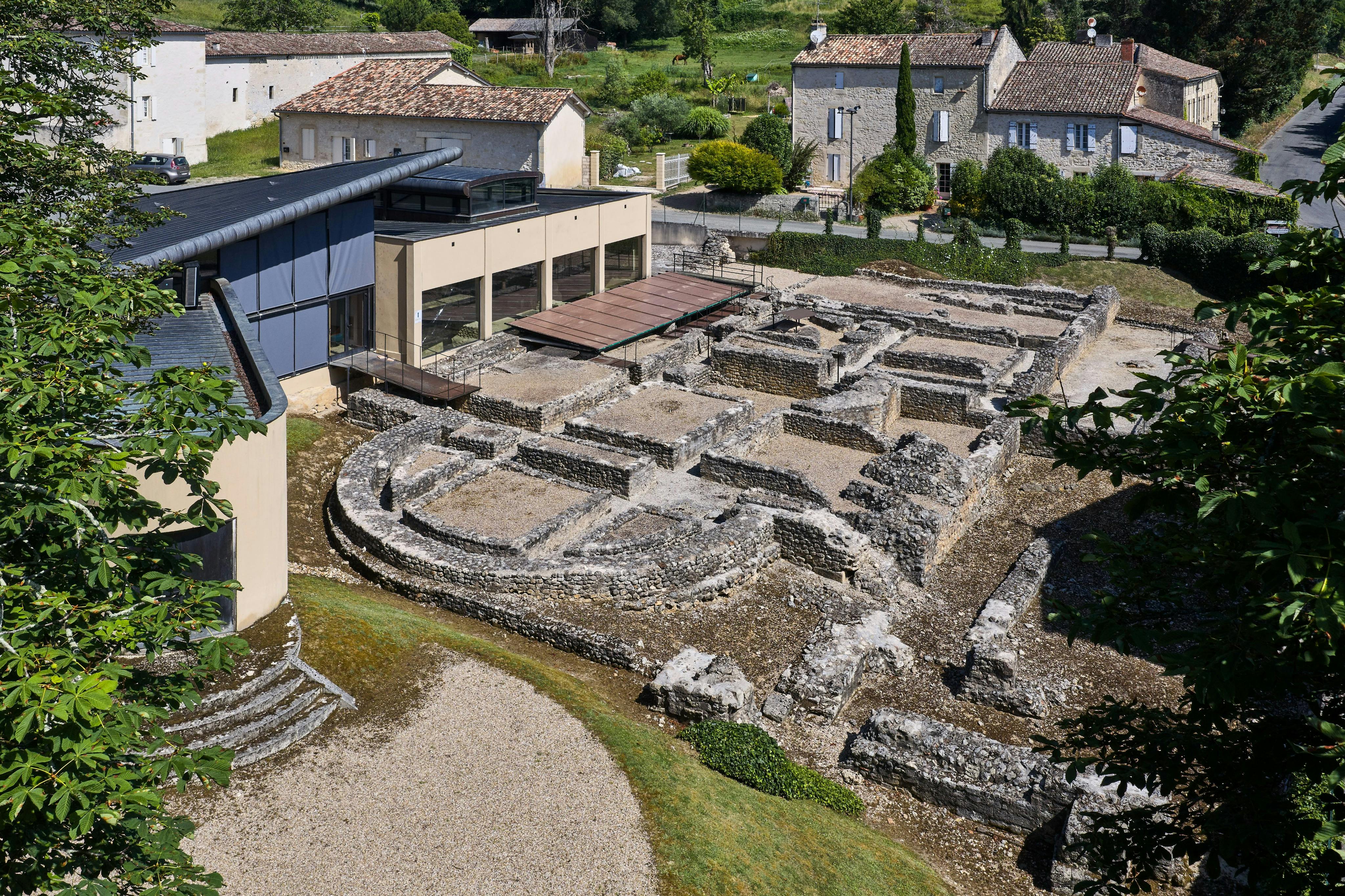 Antiguas ruinas de piedra junto a un edificio moderno, con casas antiguas y vegetación verde al fondo.