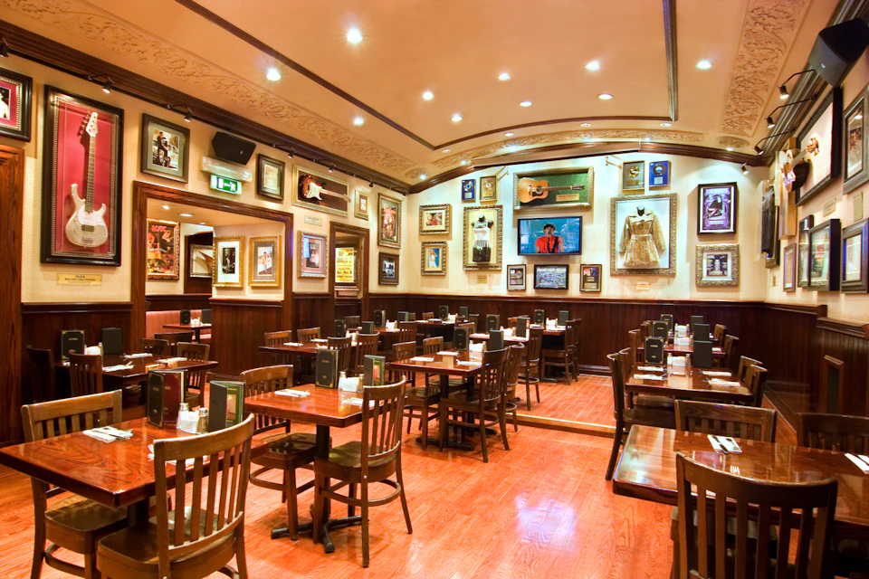 A well-lit, empty restaurant with wooden tables and chairs. Walls are adorned with framed photos, memorabilia, and a TV screen.