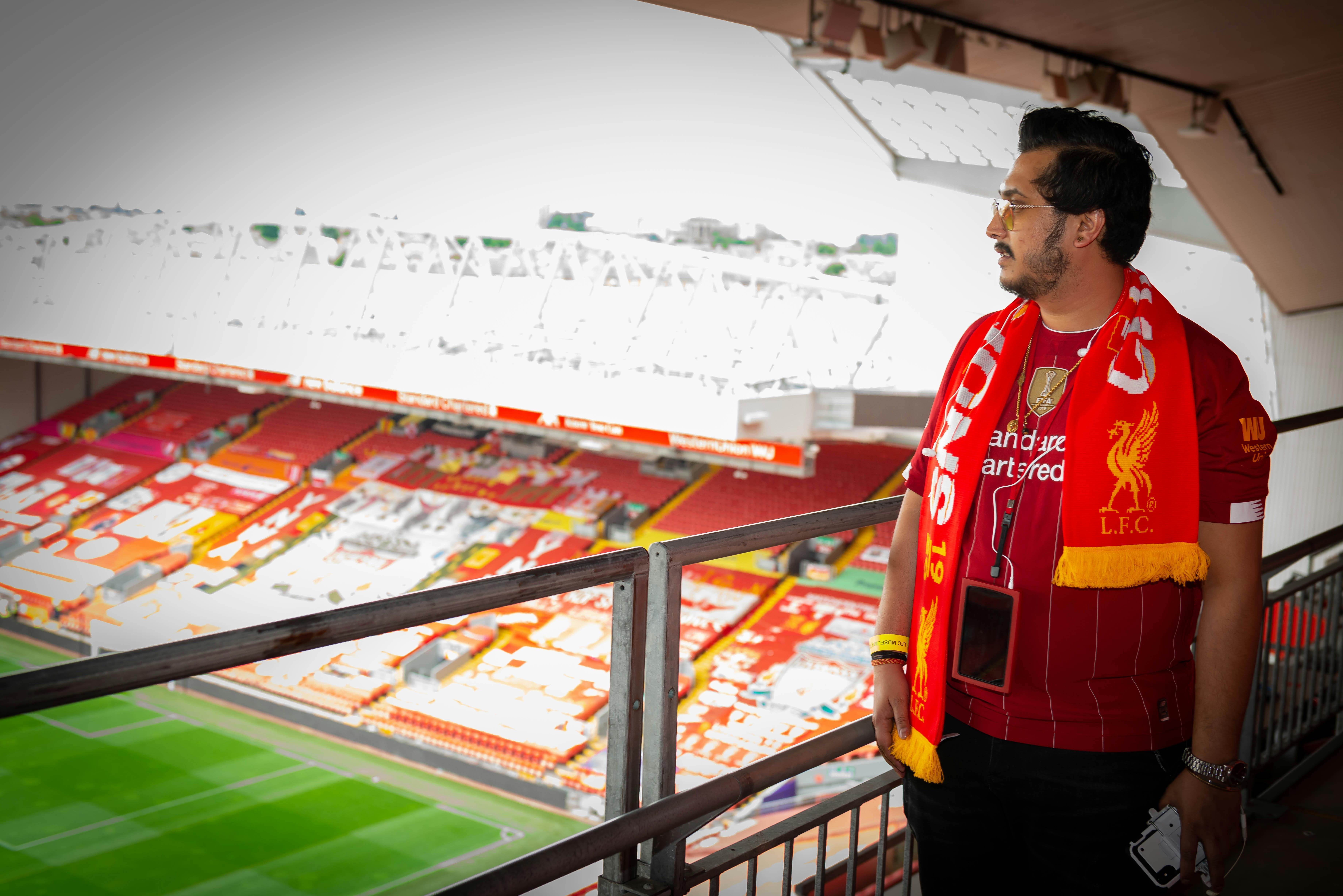 A man wearing a red LFC shirt and scarf stands in a stadium, looking out over the empty seats and pitch.
