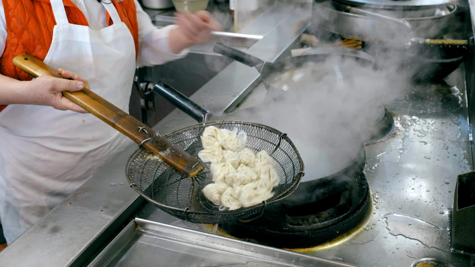 A person wearing a white apron is using a wire strainer to cook dumplings in a steaming pot in a kitchen.