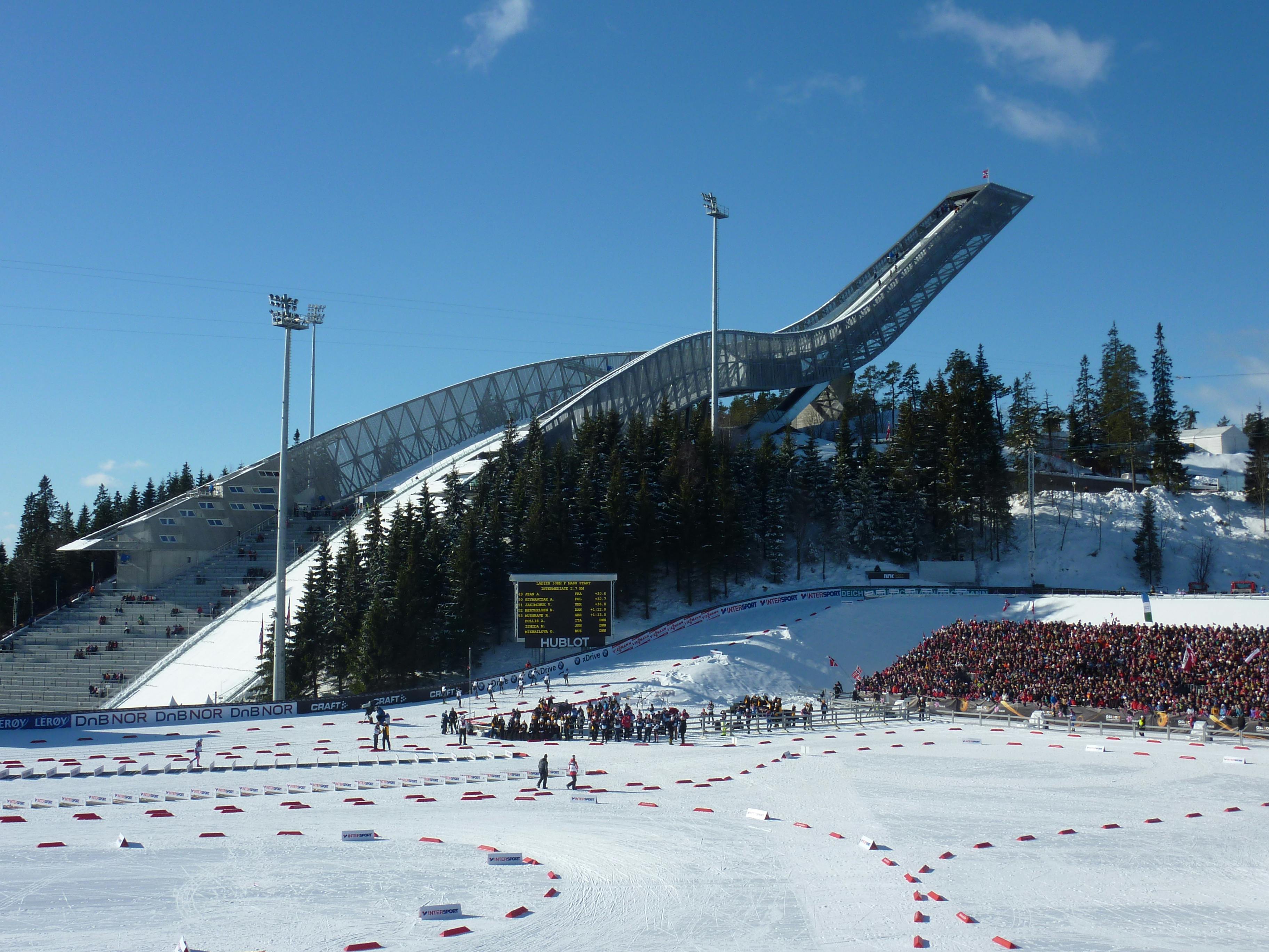 Πλήθος κόσμου παρακολουθεί μια εκδήλωση σκι με ένα μεγάλο άλμα σκι και χιονισμένο τοπίο, δέντρα και πίνακα αποτελεσμάτων στο παρασκήνιο κάτω από έναν καθαρό ουρανό.