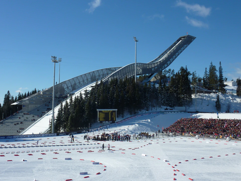 Crowd watching a ski event with a large ski jump and snowy landscape, trees, and scoreboard in the background under a clear sky.