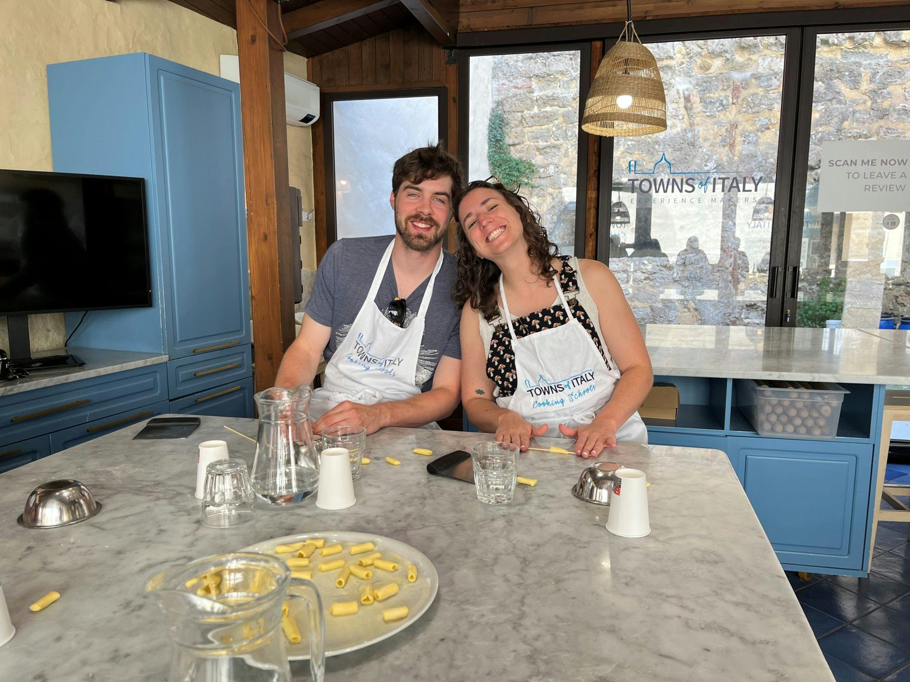 Two people smiling while making pasta at a cooking class in a kitchen with light blue cabinets and a marble countertop.
