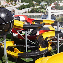 Ett kluster av färgglada vattenrutschbanor flätas samman vid en vattenpark, med pooler och grönska i bakgrunden.