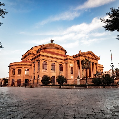 Large, historic building with a domed roof and columned entrance, surrounded by trees and a flagpole, set against a blue sky.