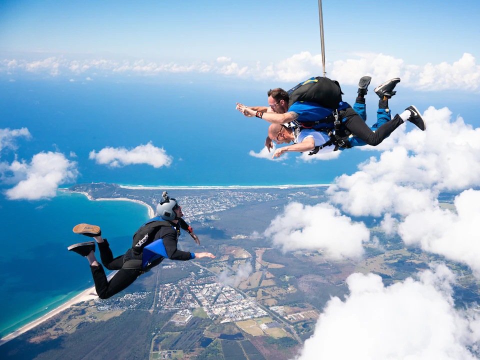 Three skydivers in mid-air above a coastal landscape with clouds, ocean, and a town visible below.