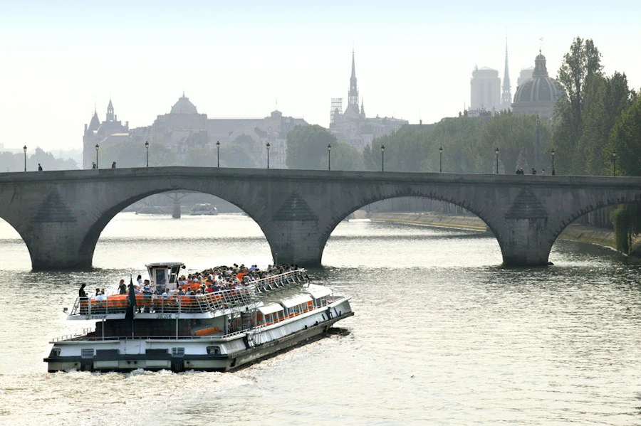 Barco lleno de pasajeros pasando bajo un puente de piedra, con el horizonte de la ciudad detrás