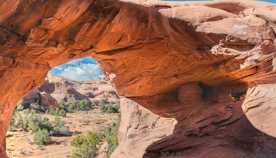 A natural red rock arch frames a desert landscape with trees, bushes, and a blue sky with clouds.