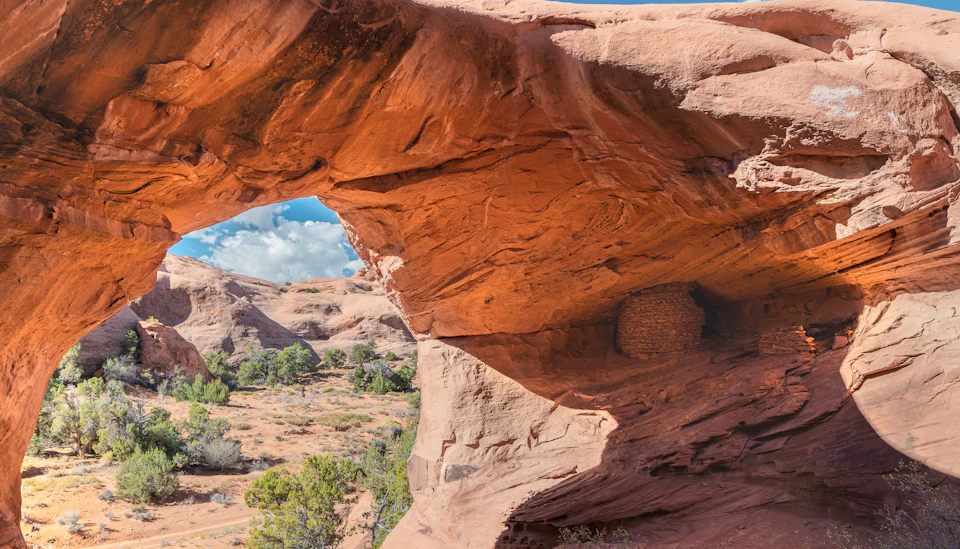 A natural red rock arch frames a desert landscape with trees, bushes, and a blue sky with clouds.