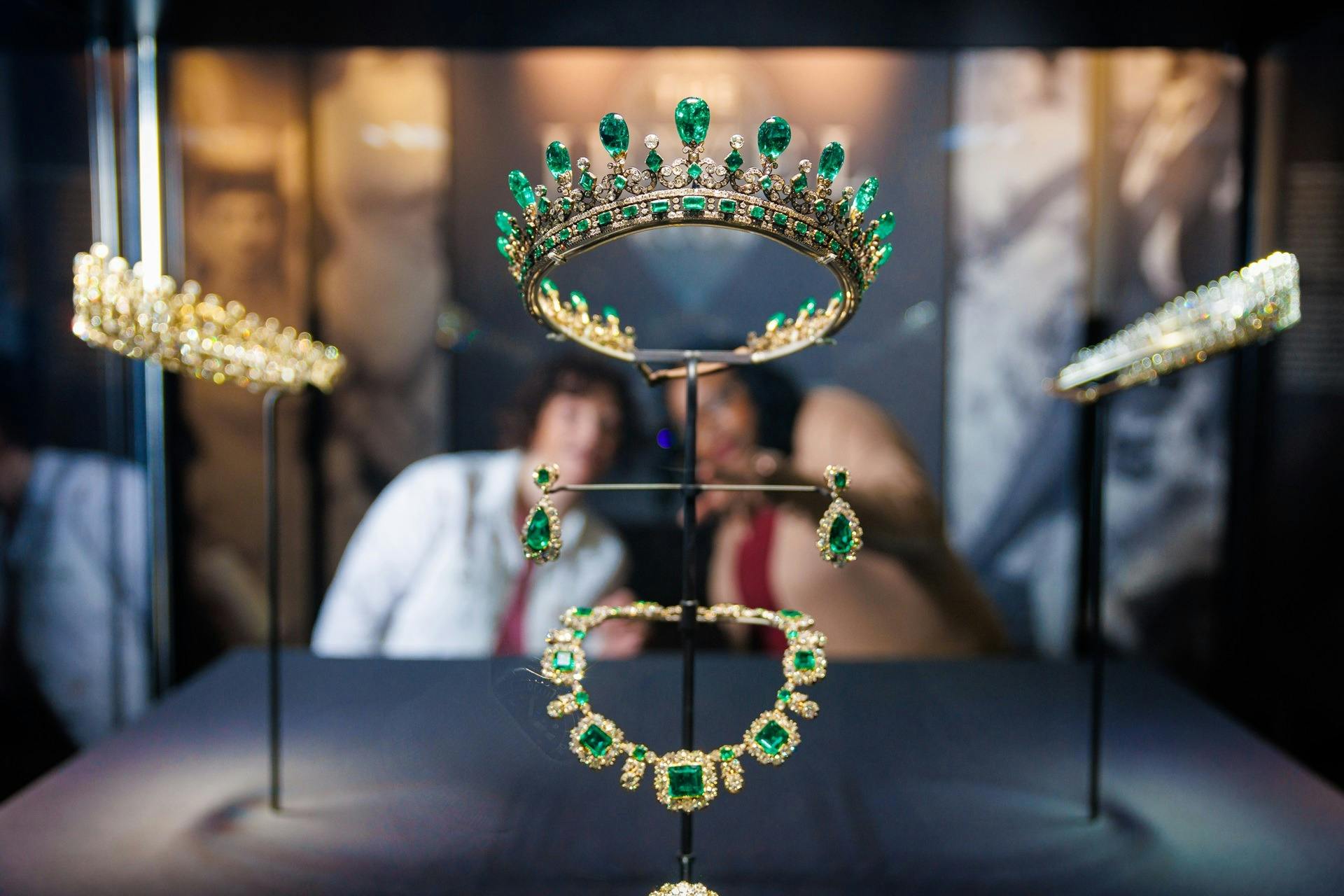 A display of emerald and gold jewelry, including a tiara, earrings, and necklace, with two blurred people in the background.