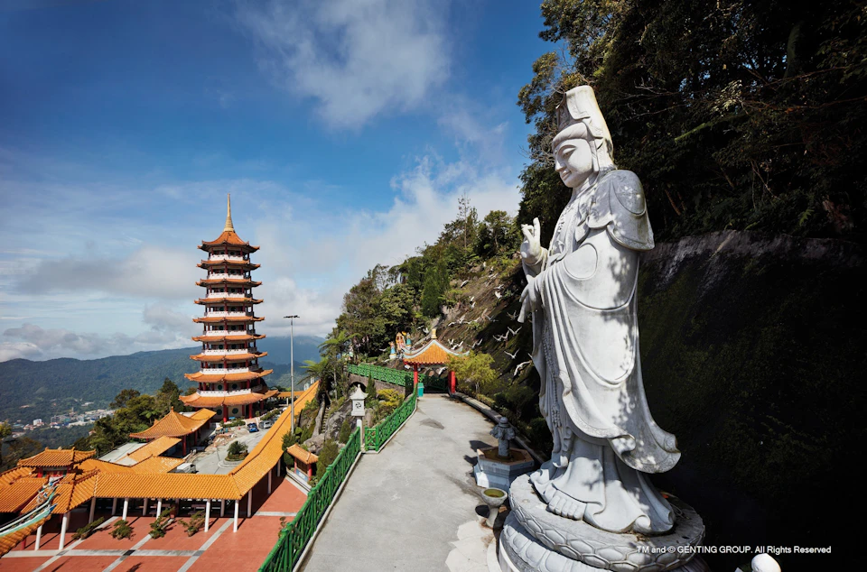 A tall pagoda and a large statue of a deity in a mountain setting, with a pathway and greenery under a blue sky.