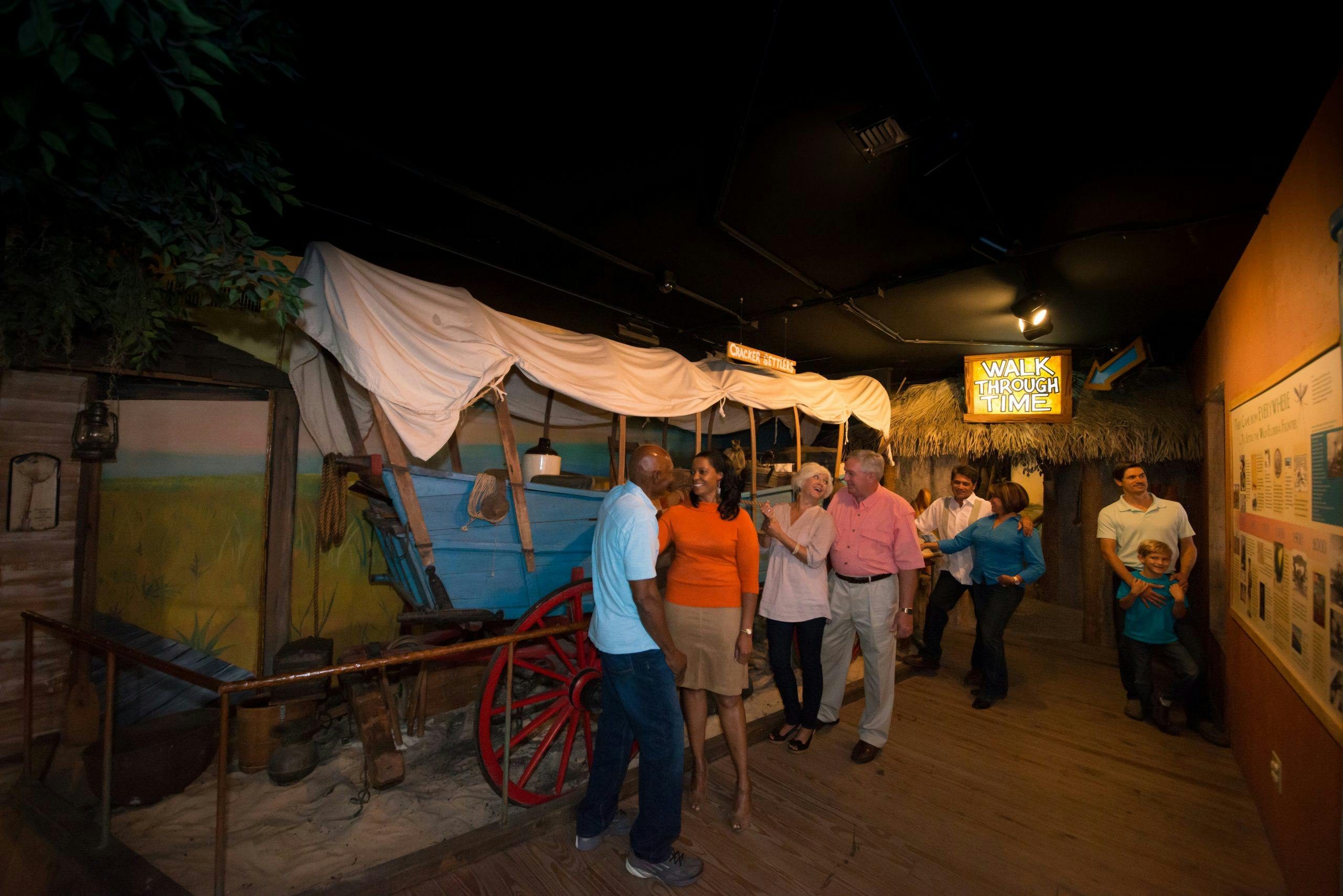 A group of people stand and talk near a covered wagon exhibit indoors under a sign that reads "WALK THROUGH TIME."