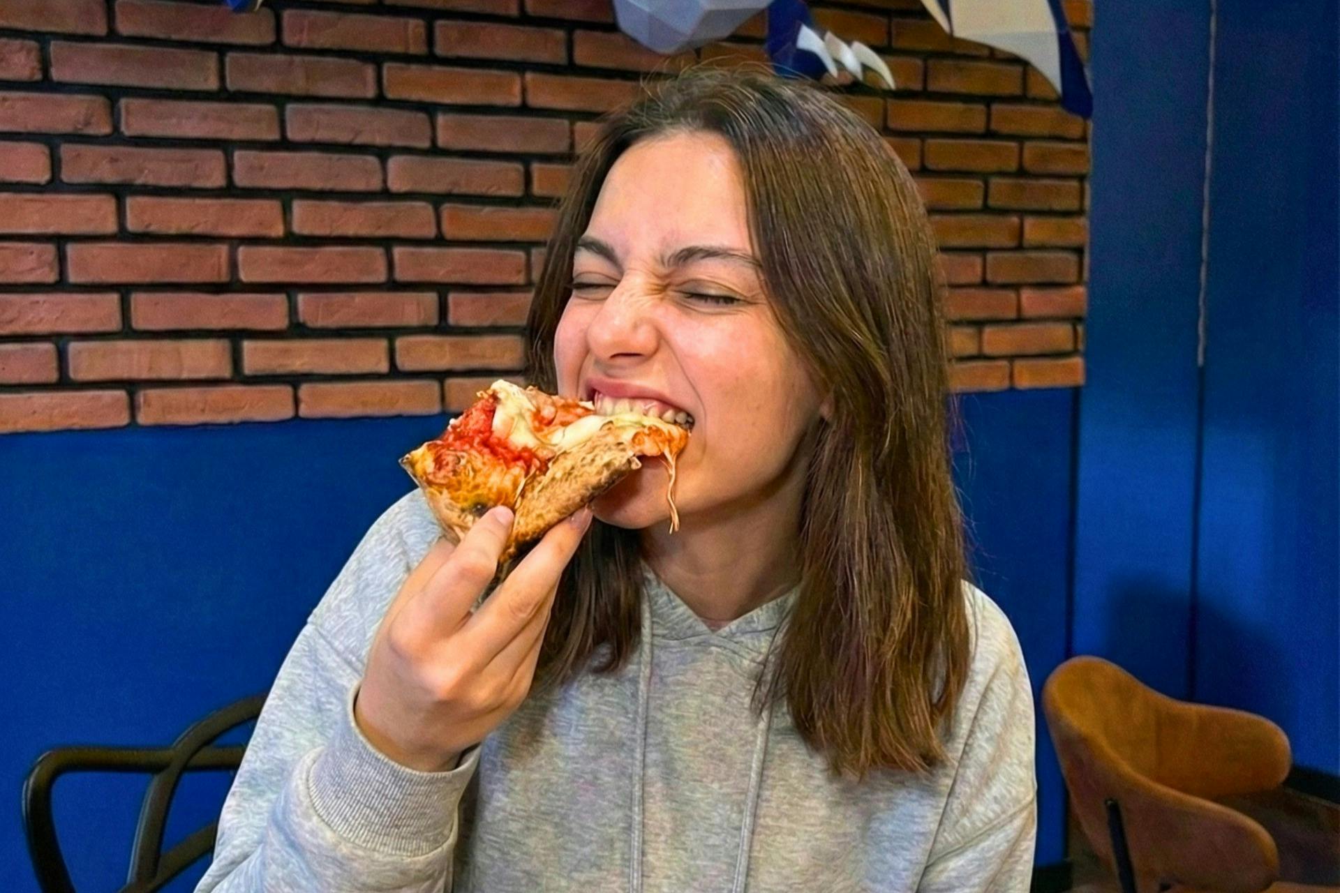 A person in a hoodie is enthusiastically eating a slice of pizza in a room with a brick wall and blue accents.
