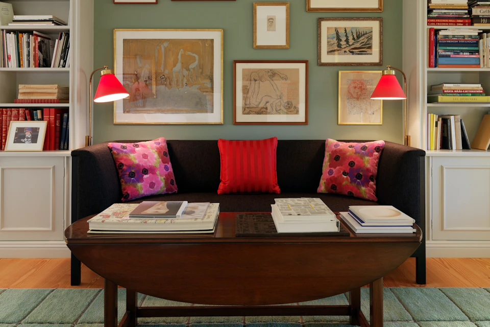 A living room with a black sofa, red and floral cushions, a wooden coffee table with books, and framed artwork on the green wall.