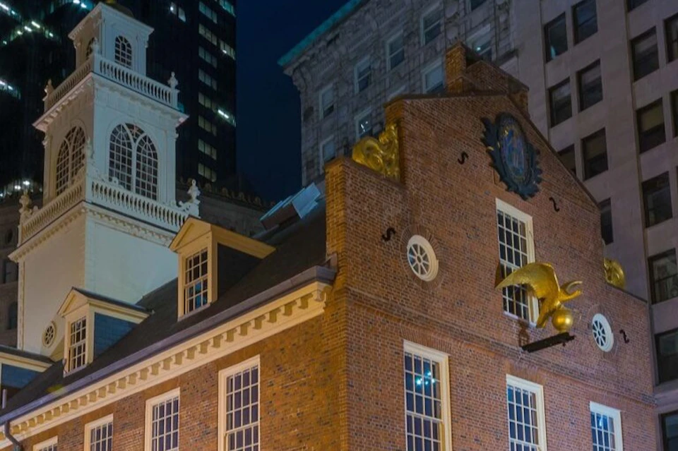 A historic red brick building with golden ornaments and a clock, surrounded by modern and classical architecture at night.