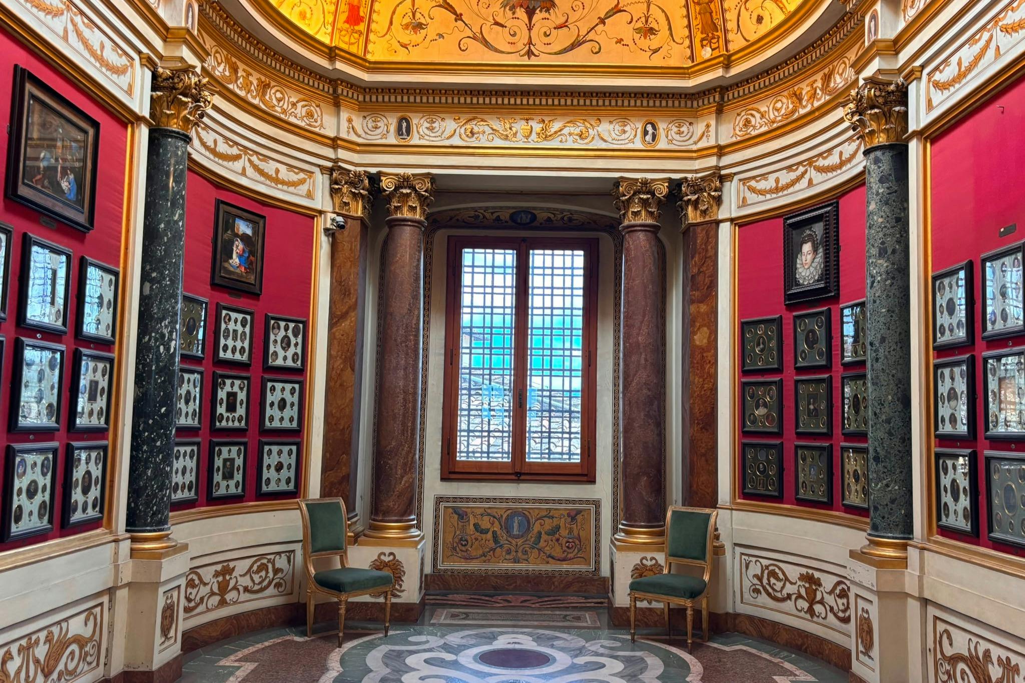 Uma sala ornamentada com um teto abobadado, colunas de mármore, retratos emoldurados nas paredes vermelhas e uma janela que dá para um céu luminoso.