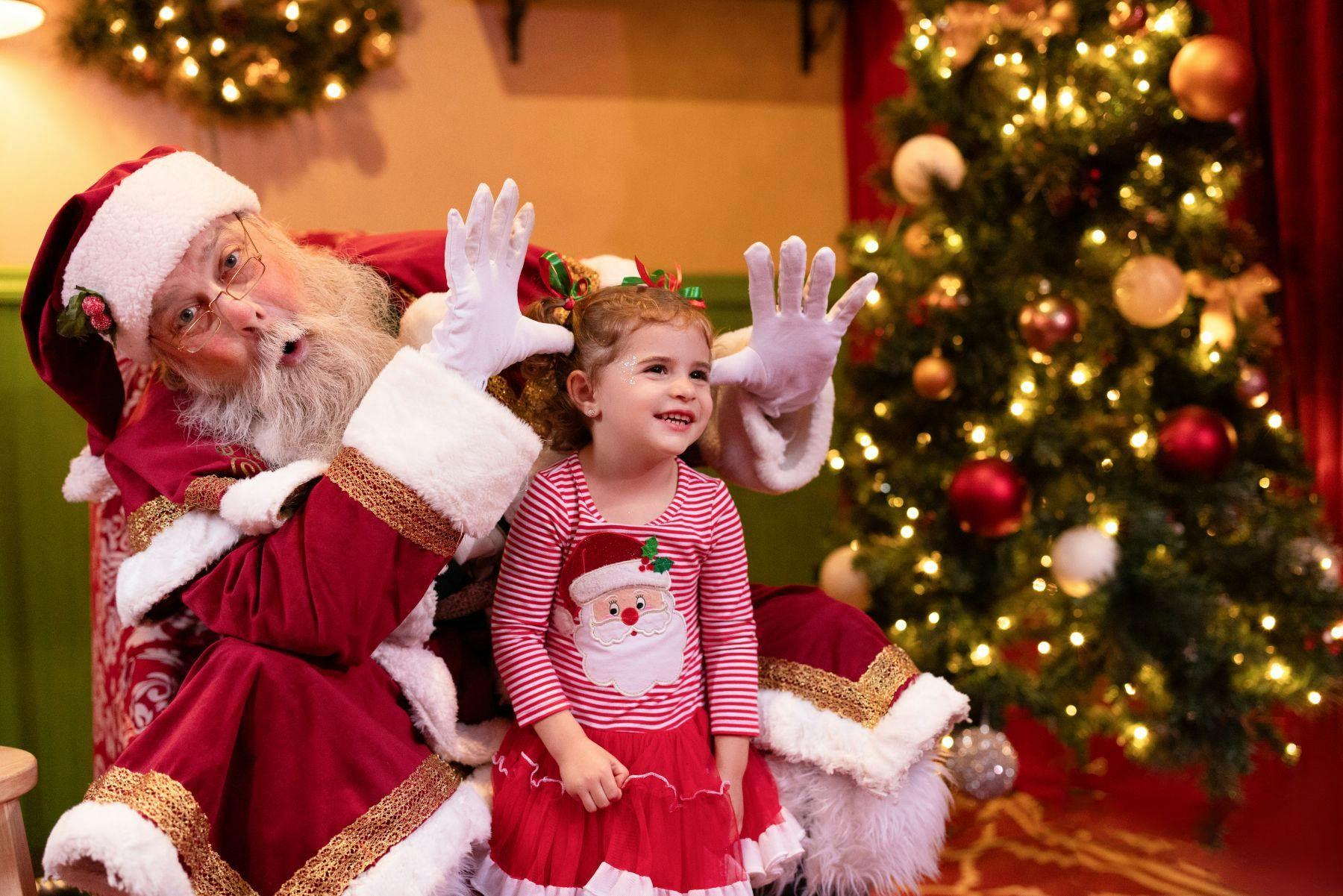 A child in a festive outfit sits with Santa Claus, smiling, in a decorated holiday setting with a Christmas tree in the background.