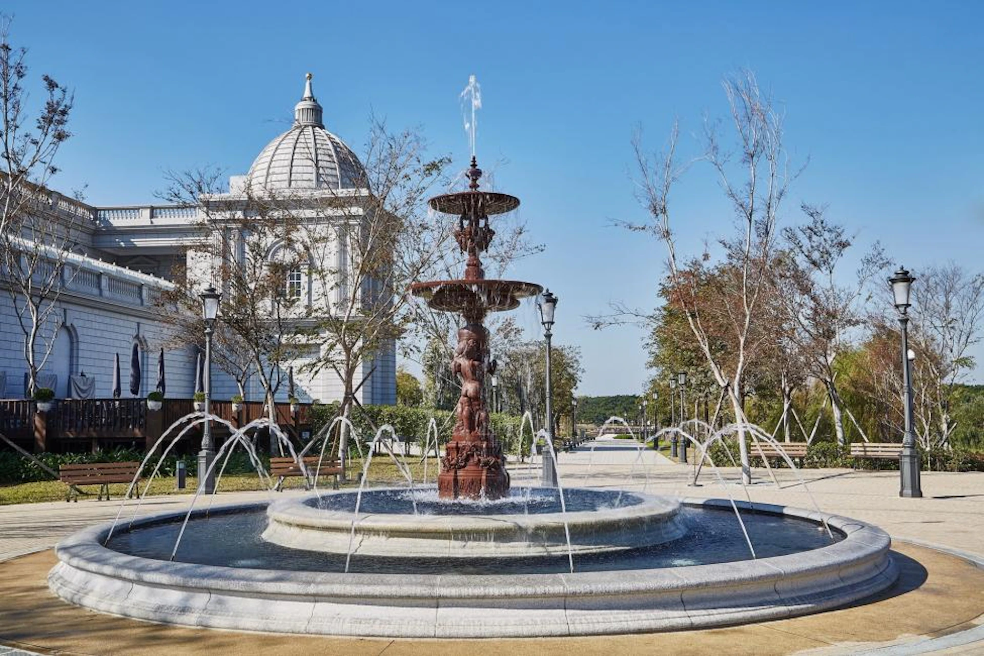 A decorative fountain with jets of water surrounded by trees and a domed building under a clear blue sky.