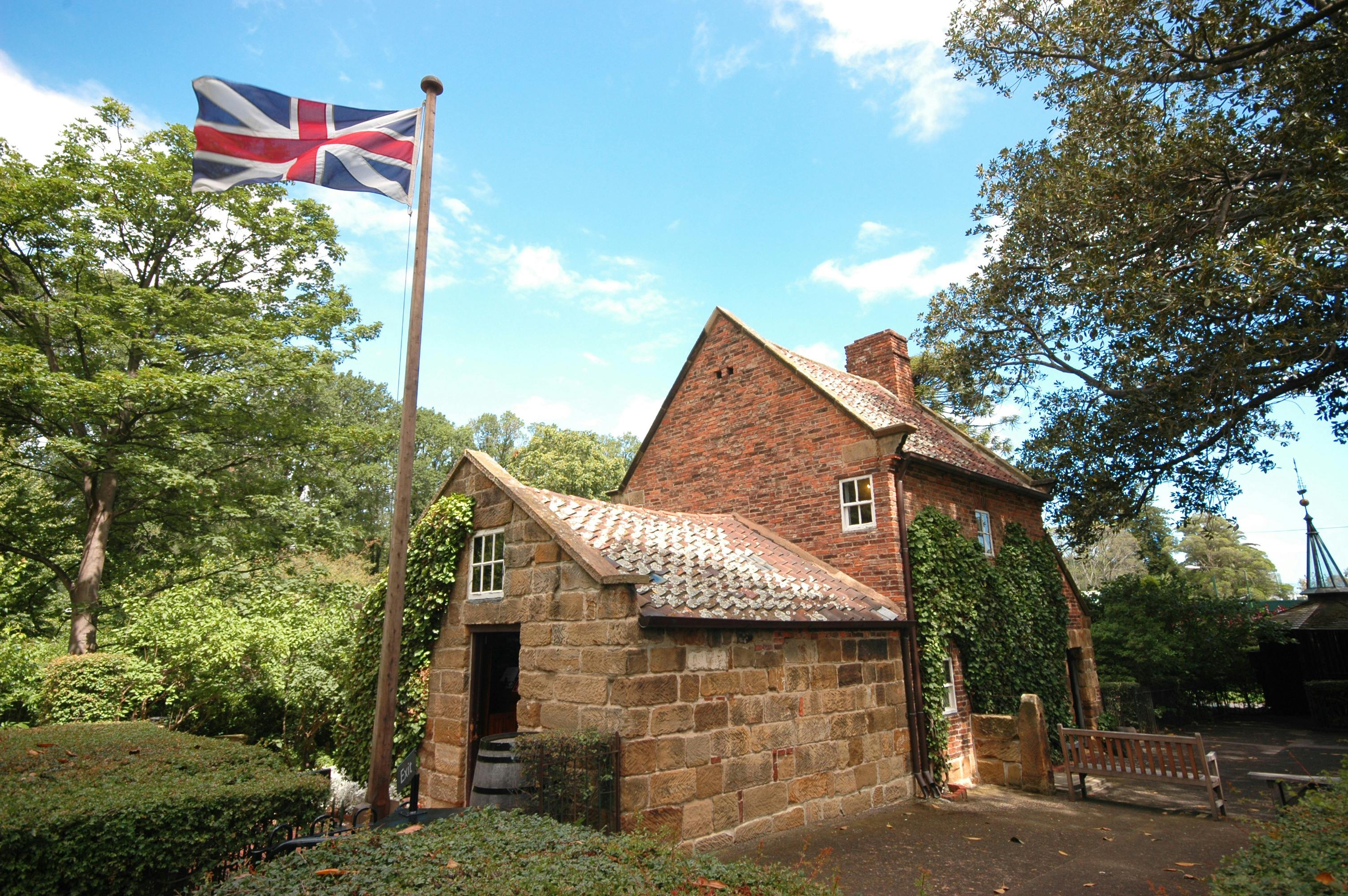 Historic brick house with ivy-covered walls, a stone extension, and a Union Jack flag in the foreground. Trees surround the building.