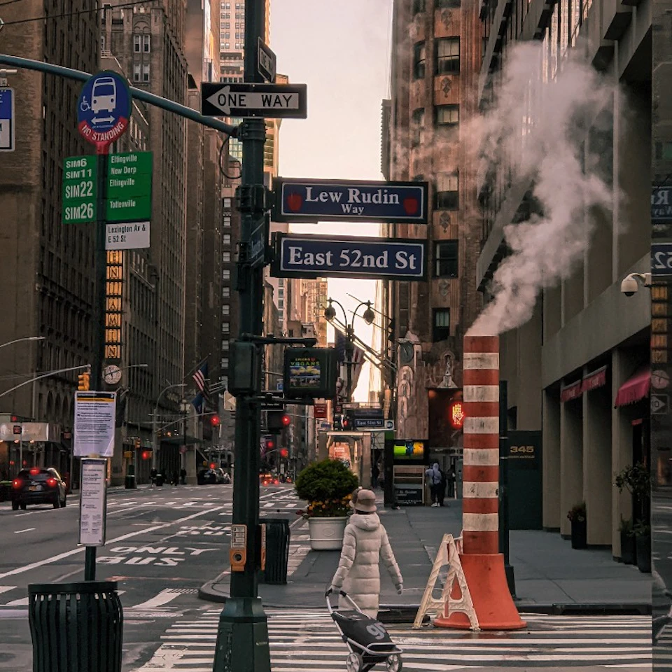 A New York City street intersection with a steam vent, street signs for "Lew Rudin Way" and "East 52nd St", and a person pushing a stroller.