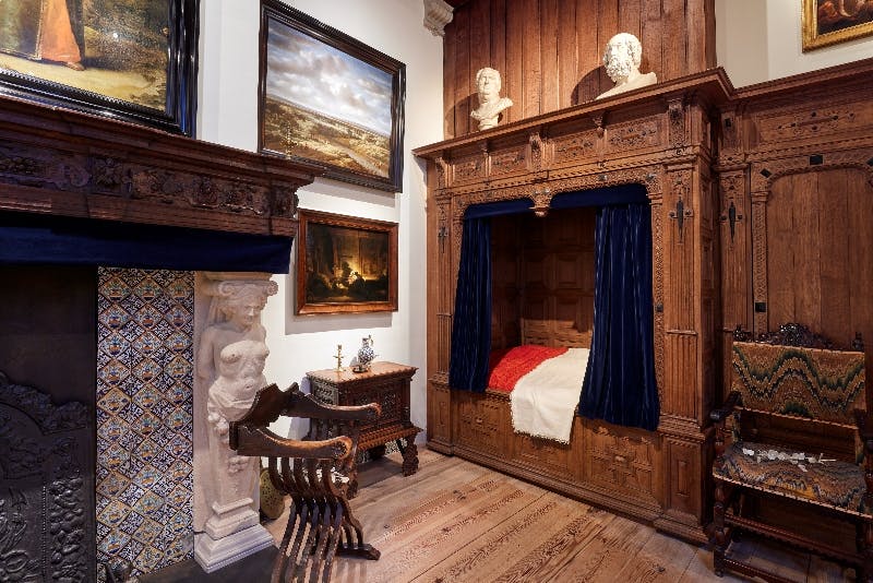 Ein historisches Zimmer mit einem hölzernen Himmelbett, blauen Vorhängen, einem verschnörkelten Kamin, Gemälden an den Wänden und antiken Möbeln.