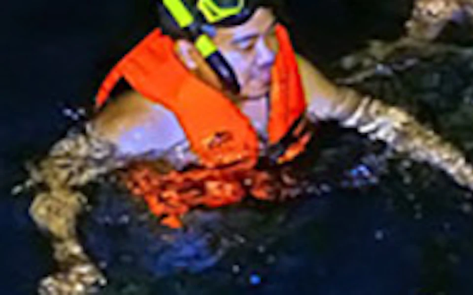 People wearing life vests and snorkeling gear float in dark water with bioluminescence creating a glowing effect around them.
