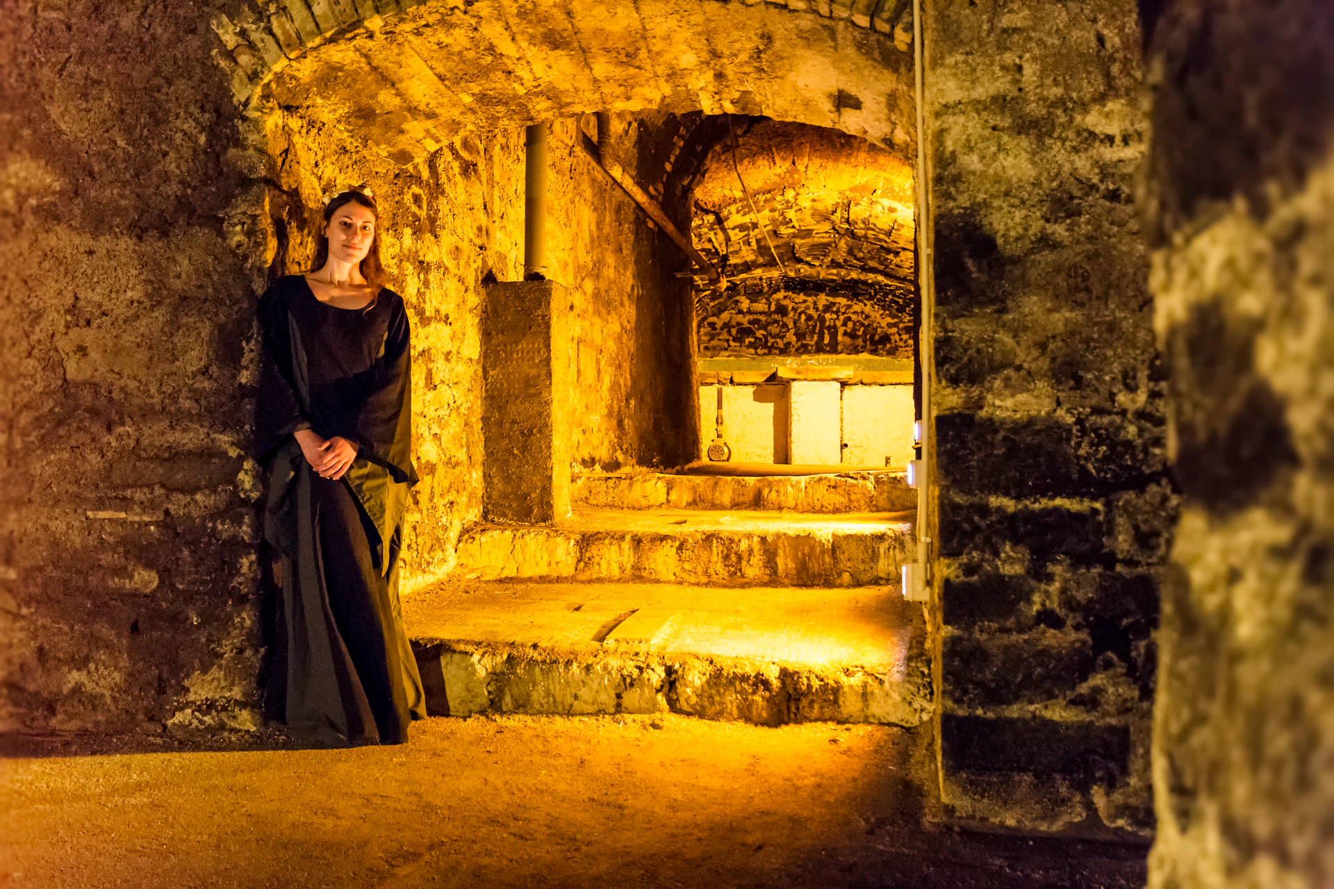 A woman in a long dark dress stands in a dimly lit stone-walled passageway with arches and a staircase illuminated in warm light.