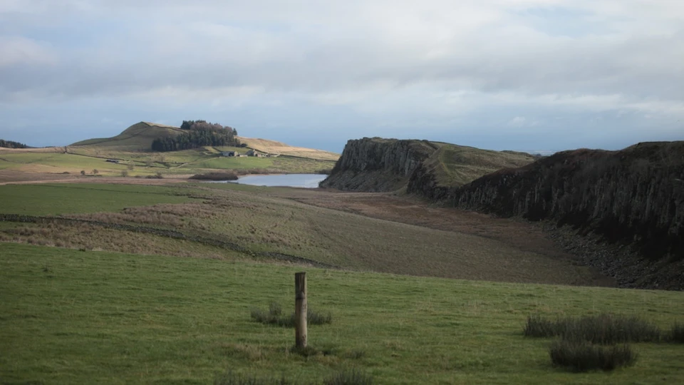 A grassy landscape with a wooden post in the foreground, rocky cliff, pond, and cloudy sky in the background.