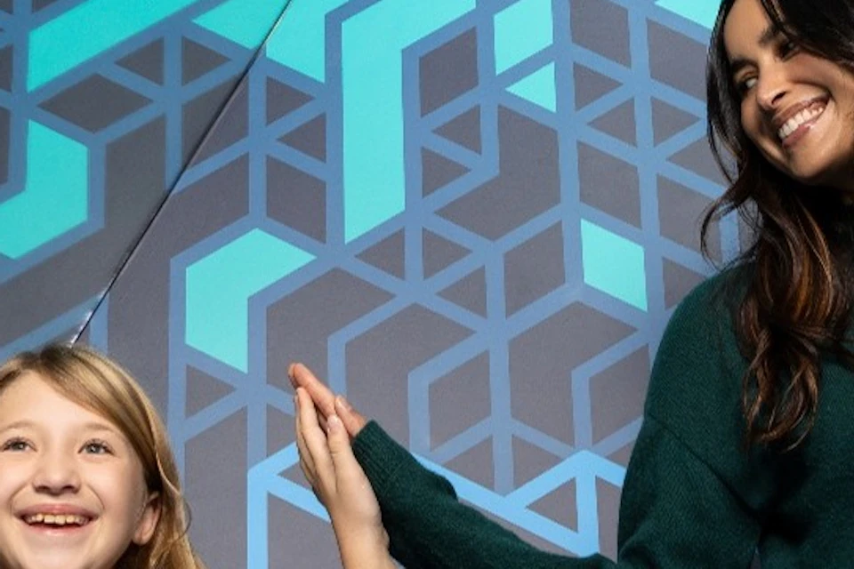 Two people, a child in a purple shirt and an adult in a green sweater, stand in front of a geometric mirrored wall.