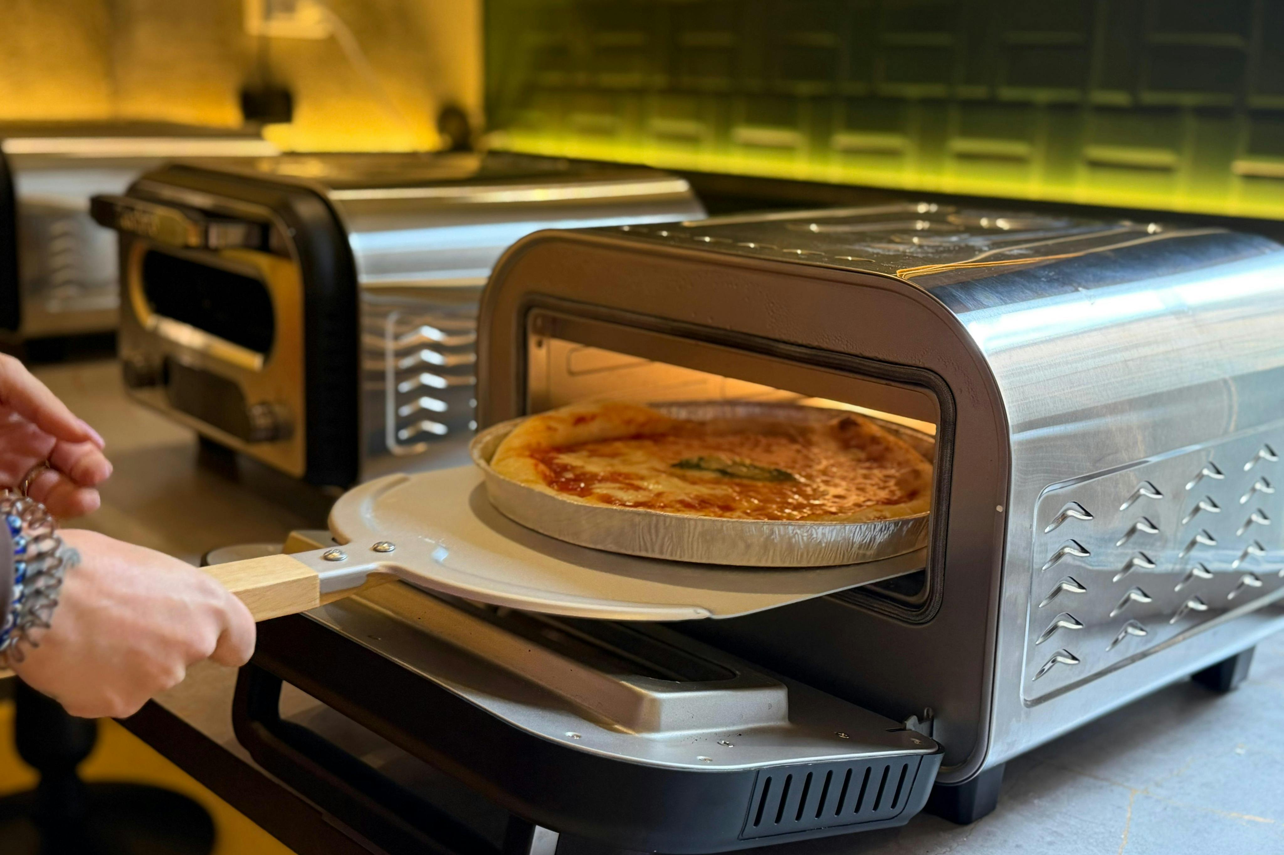 A pizza being placed into a silver countertop pizza oven using a wooden pizza peel.