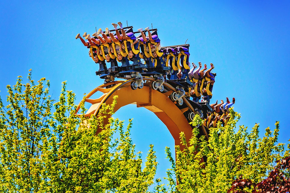 A group of friends laughing and hugging at an amusement park, with a roller coaster and American flags in the background.
