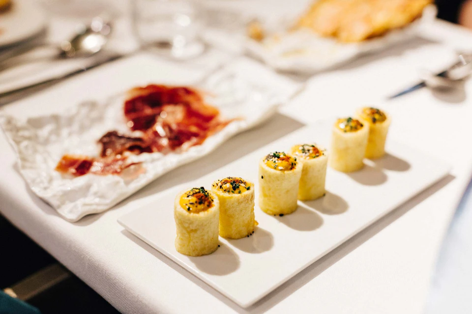 Four sushi rolls are neatly arranged on a white rectangular plate, with a plate of thinly sliced meat in the background.