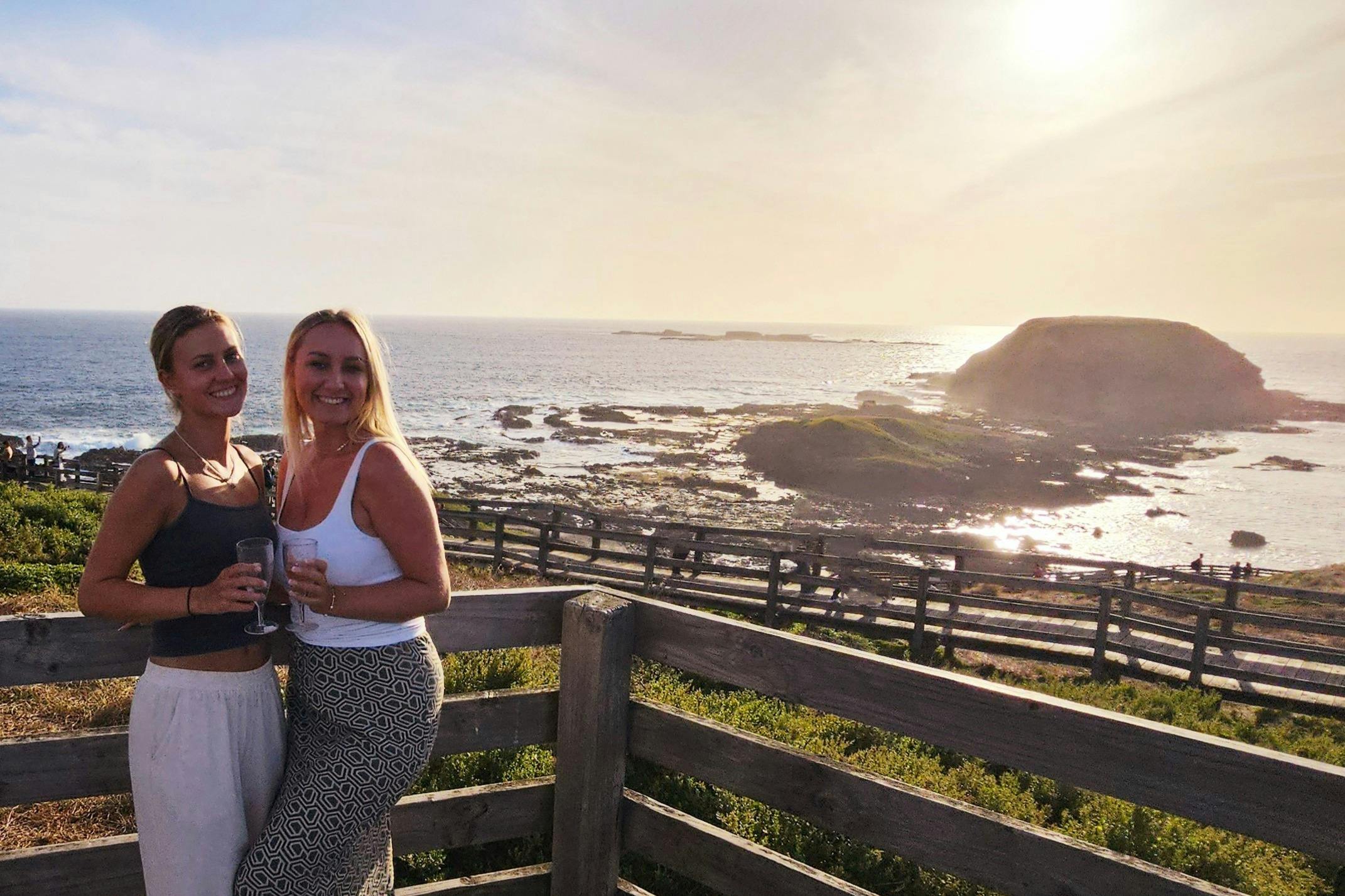 Two women smile while holding drinks, standing by a wooden railing with an ocean and rocky coastline in the background during sunset.
