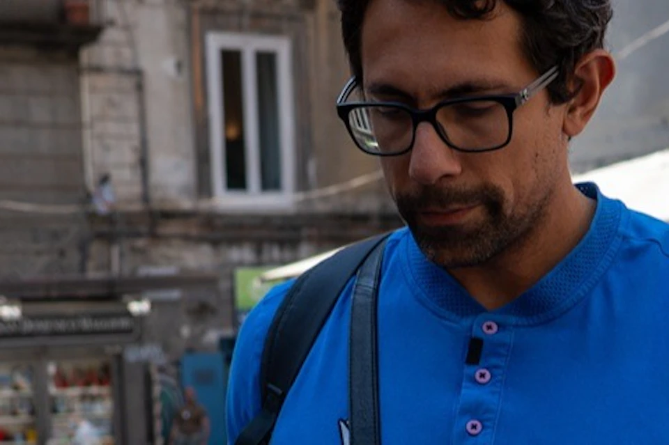Three people interacting outdoors in a sunny urban setting, with one person handing items to two others, possibly lanyards.